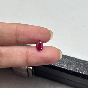 Puede incluir: Una pequeña piedra preciosa de rubí de corte ovalado con un color rojo profundo y saturado y una superficie facetada que refleja la luz. La piedra se sostiene entre dos dedos, mostrando su calidad translúcida y su acabado pulido.