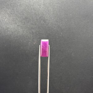 May include: A rectangular, faceted gemstone with a vibrant magenta-pink hue held by a pair of silver-toned metal tweezers. The stone features a flat top surface and angled edges that catch the light, displaying a consistent, saturated color throughout.