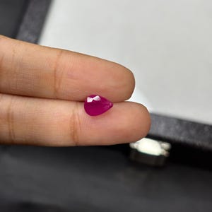 以下が含まれることがあります： ファセット加工された、洋梨型のルビーの宝石。ルビーは深く鮮やかな赤色で、2本の指でつまんでいます。宝石は研磨された表面と尖った底を持っています。背景はぼやけています。