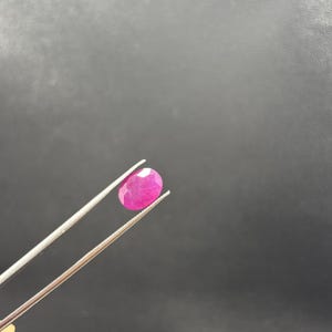 May include: A vibrant, oval-shaped, faceted pink gemstone held by a pair of silver-toned metal tweezers. The gemstone displays a translucent quality with visible internal inclusions and a polished surface, reflecting light across its many cut facets.