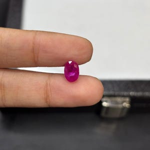 Puede incluir: Una vibrante gema de rubí de corte ovalado. El rubí es de un color rojo intenso y rico, con facetas que capturan la luz. La gema se sostiene entre dos dedos, mostrando su tamaño y brillo. El fondo es blanco y negro neutro.