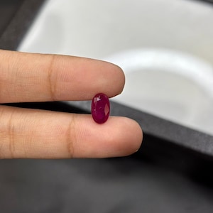 May include: A faceted, oval-shaped ruby gemstone. The ruby is a deep, rich red color. The gemstone is held between two fingers, showcasing its polished surface and brilliance. The background is a neutral gray.