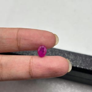Könnte beinhalten: Ein kleiner, oval geschliffener, leuchtend pink-roter Edelstein mit einer facettierten Oberfläche, die das Licht reflektiert. Der Edelstein wird zwischen zwei Fingern gehalten und zeigt seine durchscheinende Beschaffenheit sowie seine tiefe, gesättigte Farbe.