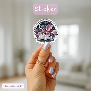 Könnte beinhalten: Ein Aufkleber mit Fantasy-Motiv. Er zeigt einen rosa Drachen auf einem offenen Buch, umgeben von Blumen und dem Text "Fantasy Readers Book Club". Das Wort "sticker" steht oben. Der Aufkleber wird von einer Hand mit schillernden Nägeln gehalten.