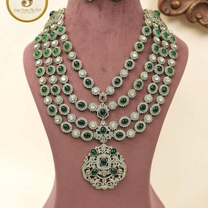 May include: A multi-strand necklace set with matching earrings and a brooch. The jewelry features green and clear gemstones set in a silver-toned metal. The necklace has a layered design, and the earrings are round studs. The brooch is a detailed pendant.