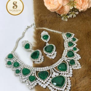 May include: A silver-toned necklace and matching drop earring set featuring large, faceted, heart-shaped emerald green stones surrounded by small, clear, sparkling white crystals in a decorative, intricate metal setting with a chain.
