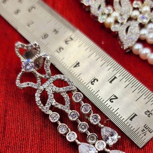 May include: Silver dangle earrings with a teardrop design and clear cubic zirconia stones. The earrings are measured against a ruler.