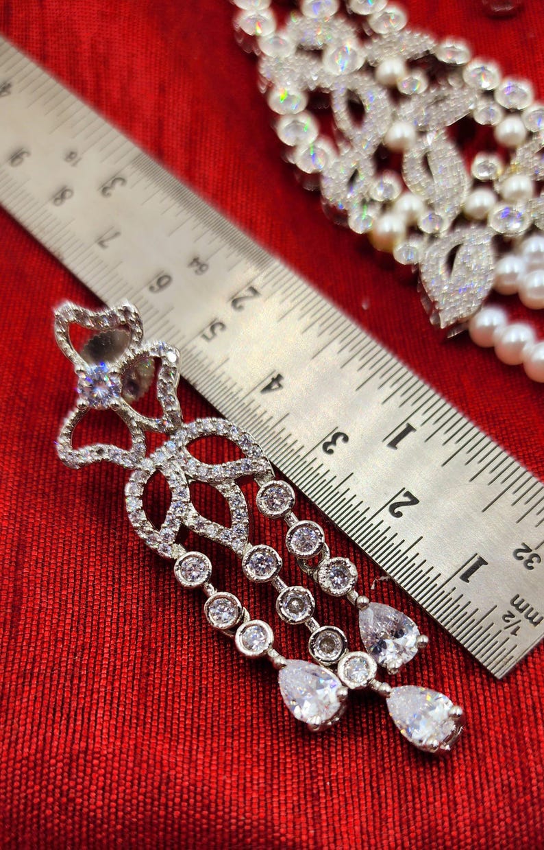 May include: Silver dangle earrings with a teardrop design and clear cubic zirconia stones. The earrings are measured against a ruler.