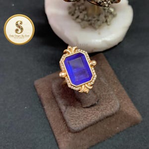 May include: A gold ring featuring a large, rectangular, deep blue gemstone set in the center. The stone is surrounded by a border of small, clear, sparkling stones. The ring has a decorative gold metal frame with small spheres at the corners.