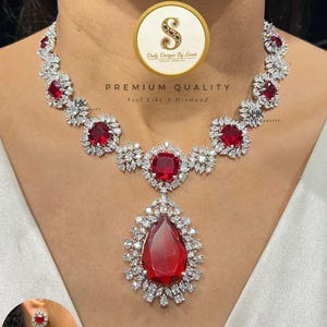 Op de afbeelding: Een schitterende ketting en bijpassende oorbellen set. De ketting heeft rode edelstenen omringd door heldere, sprankelende stenen. De hanger is een grote, druppelvormige rode edelsteen. De tekst luidt "Premium Quality, Feel Like a Diamond."