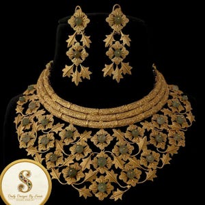 May include: A gold-toned necklace and earring set. The necklace features a floral design with green gemstones and leaf accents. The earrings have a similar design, with dangling leaf and floral elements. The jewelry is displayed on a black background.