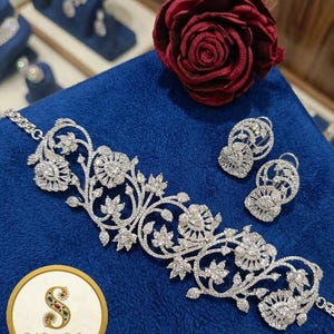 Può includere: Un set di gioielli argentati composto da un braccialetto e orecchini abbinati, ornati di pietre preziose trasparenti. Il set è presentato su una superficie blu, con una rosa rossa sullo sfondo. È visibile anche il logo Daily Designs By Swati.