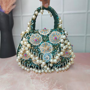 Peut inclure: Un sac à main vert foncé de forme arrondie, orné de perles blanches et de broderies complexes. Le sac est doté d'une poignée et est orné de paillettes et de perles dorées, bleues et roses. Le design est orné et élégant.