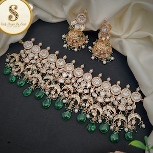 Peut inclure: Un ensemble de bijoux indiens de couleur dorée, comprenant un collier et des boucles d'oreilles assorties. Le collier est orné de pierres rondes transparentes et d'éléments verts en forme de goutte. Les boucles d'oreilles ont un design similaire. L'ensemble est présenté sur une surface noire.