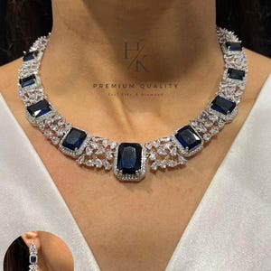 Puede incluir: Un deslumbrante conjunto de collar y pendientes con grandes piedras preciosas rectangulares de color azul oscuro engastadas en metal plateado. El collar tiene un diseño simétrico con piedras más pequeñas y claras. El texto "PREMIUM QUALITY" es visible.