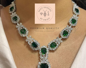 May include: A dazzling necklace featuring emerald-cut green gemstones and sparkling clear stones. The necklace is set in a silver-toned metal and is displayed on a person. The image includes the text "DAILY DESIGNS BY SWATI" and "PREMIUM QUALITY Feel Like A Diamond."