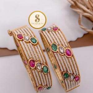 Pode incluir: Um par de pulseiras douradas com fileiras de pequenas contas brancas e pedras ovais alternadas em vermelho, verde e transparente. As pulseiras têm uma borda dourada texturizada e um logotipo circular com o texto Daily Designs By Sauti.