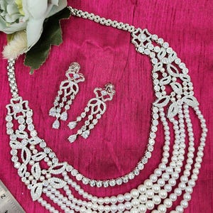 May include: A silver necklace with a multi-layered pearl strand and a floral design with clear stones. Matching earrings with a floral design and clear stones are also included.