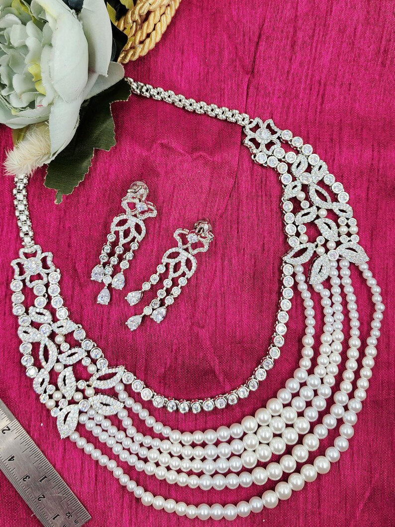 May include: A silver necklace with a multi-layered pearl strand and a floral design with clear stones. Matching earrings with a floral design and clear stones are also included.