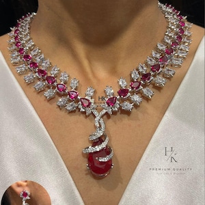 May include: A silver-toned necklace and matching earring set featuring ruby-red and clear gemstones. The necklace has a floral design with a large teardrop-shaped pendant. The earrings have a spiral design. The text "HK PREMIUM QUALITY" is visible.