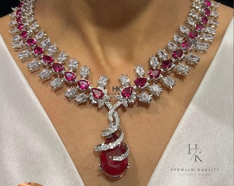 May include: A silver-toned necklace and matching earring set featuring ruby-red and clear gemstones. The necklace has a floral design with a large teardrop-shaped pendant. The earrings have a spiral design. The text "HK PREMIUM QUALITY" is visible.