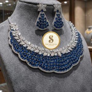 Könnte beinhalten: Ein silberfarbenes Halsketten- und Ohrring-Set mit blauen und klaren Edelsteinen. Die Halskette hat ein Blattdesign mit blauen Steinen in der Mitte. Die Ohrringe sind tropfenförmig mit blauen Steinen. Das Set wird auf einem grauen Schaufensterkopf präsentiert.