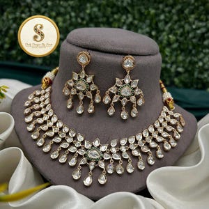 May include: A gold-toned necklace and earring set with clear, teardrop-shaped stones and green accents. The necklace and earrings are displayed on a gray bust. The image includes a logo that says "Daily Designs By Swati Indian Jewelry."