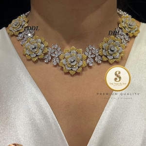 Op de afbeelding: Een bloemenketting met gele en zilverkleurige accenten. De ketting heeft een reeks bloemvormige hangers met ingewikkelde details. Er zijn ook bijpassende oorbellen te zien. De ketting is gemarkeerd met "DDL" en "Premium Quality".