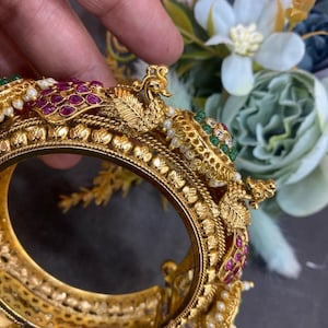 Pode incluir: Um bracelete de ouro com intrincados desenhos de pavão, adornado com pedras preciosas vermelhas e verdes e pérolas brancas.