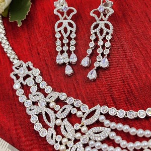 May include: A silver necklace with a pearl and crystal design. The necklace has a delicate, floral pattern and is accented with small, white pearls. The necklace is paired with matching earrings that have a similar design.