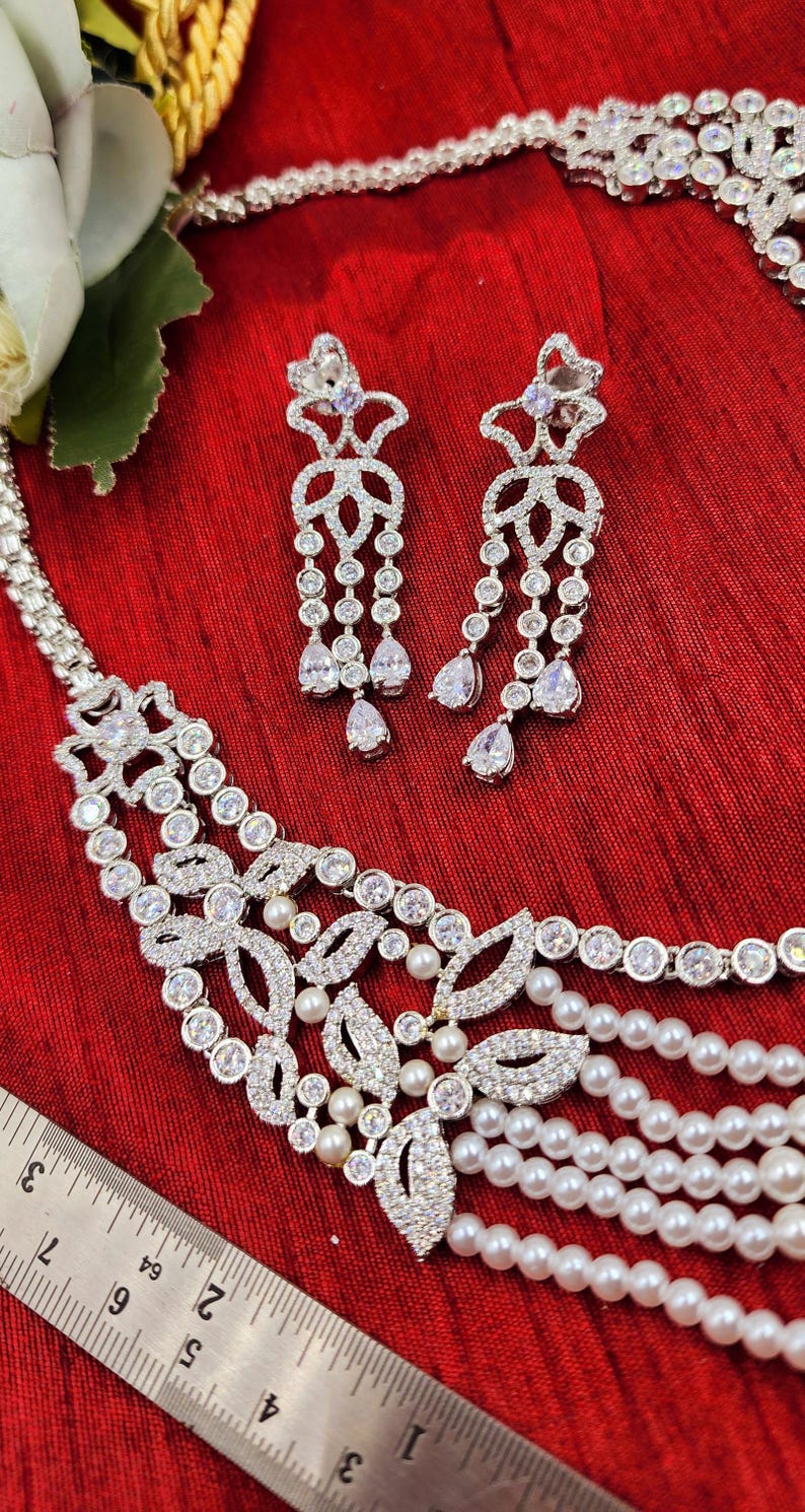 May include: A silver necklace with a pearl and crystal design. The necklace has a delicate, floral pattern and is accented with small, white pearls. The necklace is paired with matching earrings that have a similar design.