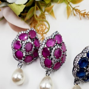 Puede incluir: Primer plano de dos pendientes ornamentados con un grupo de piedras ovaladas de color magenta rodeadas de pequeños cristales transparentes. Cada pendiente tiene una gran gota de perla blanca en forma de pera. Los pendientes están sobre un fondo blanco.