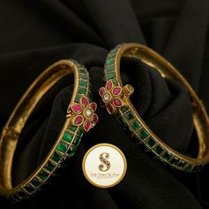 May include: Two gold-toned hinged bangles featuring a row of square-cut green stones and a central floral motif with pink petals and a clear centre stone. A circular logo with the letter S and the text Daily Designs By Swati is visible.