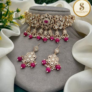 Könnte beinhalten: Ein goldfarbenes Choker-Halsband und passende Ohrhänger mit filigranen Metallarbeiten, klaren Steinen und kleinen weißen Perlen. Der Schmuck ist mit leuchtend fuchsiafarbenen Perlen akzentuiert, die an den unteren Rändern hängen.