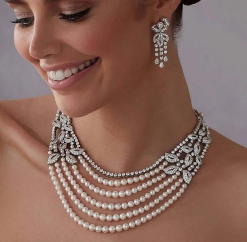 May include: A multi-strand pearl necklace with a diamond and pearl pendant. The necklace is made of white pearls and has a silver-toned metal clasp.
