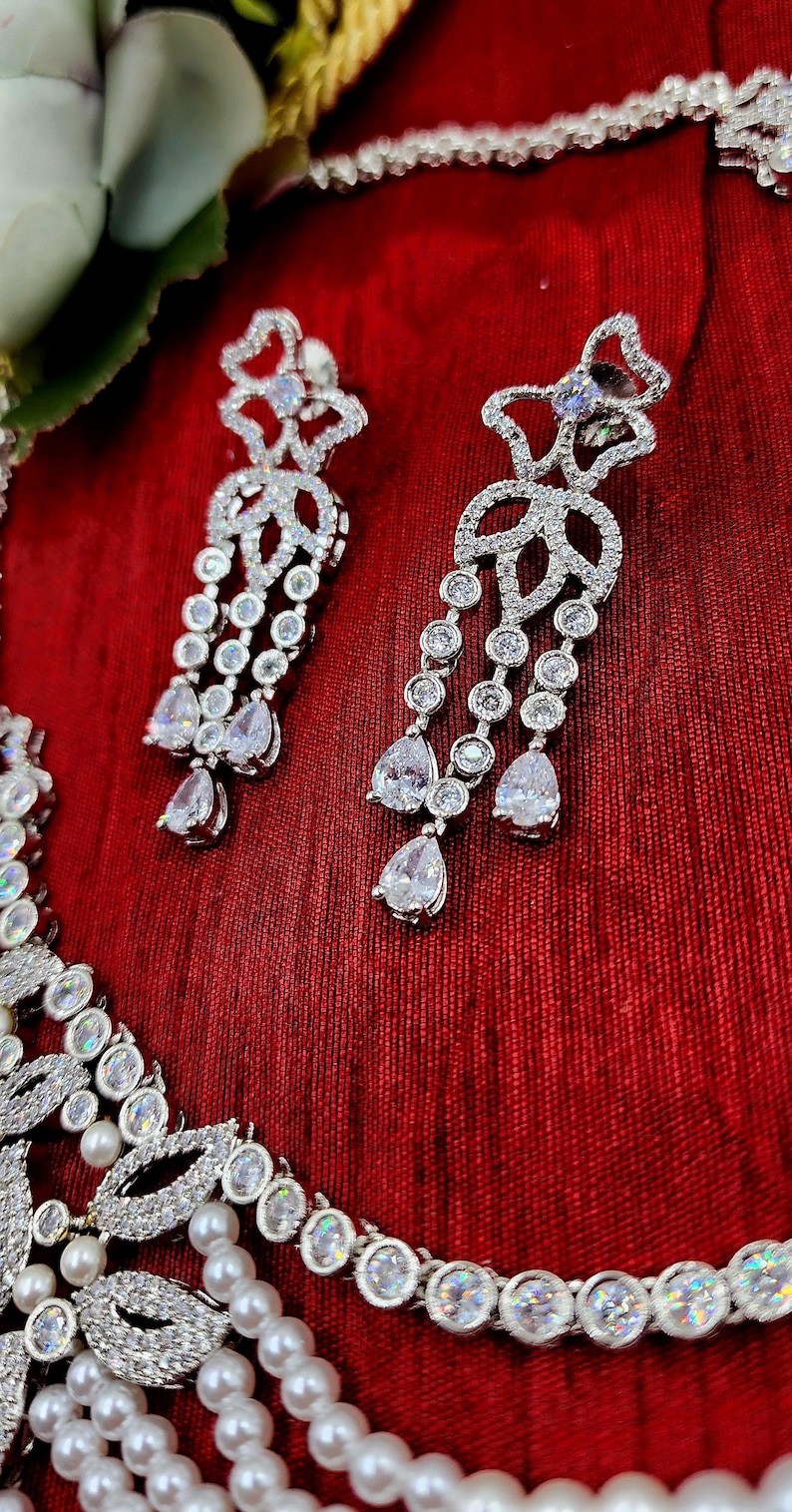 May include: A silver necklace with a pearl and crystal design, paired with matching crystal earrings. The necklace features a floral motif and the earrings are long and dangle.