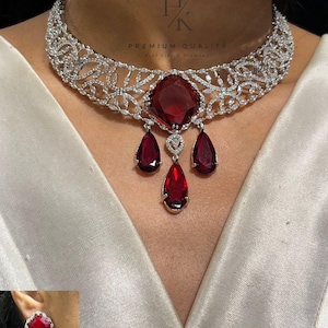 May include: A silver necklace featuring a large, square, red gemstone pendant and three teardrop-shaped red gemstones. The necklace has a floral design and matching earrings.