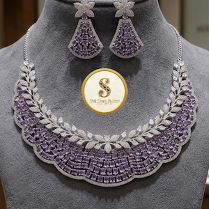 May include: A silver necklace and earring set with a floral design. The necklace and earrings feature purple and clear stones. The jewellery is displayed on a grey mannequin head.