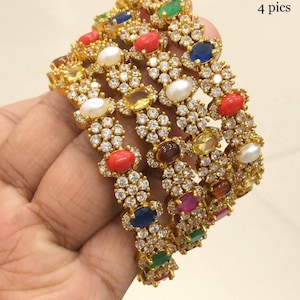 Op de afbeelding: Vier goudkleurige armbanden versierd met kleurrijke edelstenen en kleine, heldere kristallen. De ovale edelstenen zijn onder meer rood, blauw, groen, geel en wit. De tekst "4 pics" is zichtbaar.