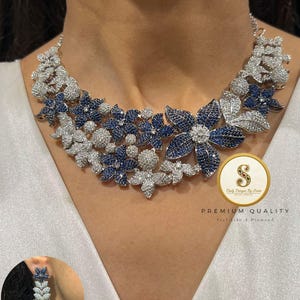 Op de afbeelding: Een bloemenketting en bijpassende oorbellen. De ketting heeft een ontwerp van blauwe en witte bloemen. De oorbellen zijn een kleinere versie van het kettingontwerp. De afbeelding bevat de tekst "PREMIUM QUALITY" en "Feel Like A Diamond".