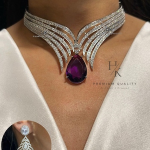 Peut inclure: Un collier et des boucles d'oreilles assorties argentés et étincelants. Le collier présente un large ras du cou avec un pendentif central en forme de goutte, orné d'une pierre précieuse violet foncé. Les boucles d'oreilles reprennent le design du collier. Le texte "PREMIUM QUALITY" est visible.