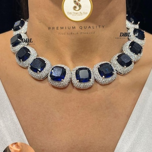 Puede incluir: Un collar lujoso con grandes piedras preciosas cuadradas de color azul oscuro engastadas en un halo de piedras más pequeñas y brillantes. El collar se lleva alrededor del cuello. También se muestra un pendiente a juego. El texto dice "PREMIUM QUALITY".