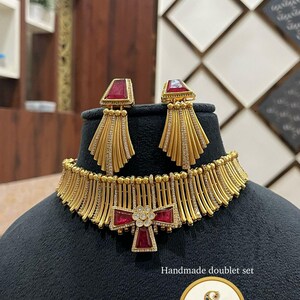 Op de afbeelding: Een goudkleurige ketting- en oorbellenset met rode edelsteenaccenten. De ketting heeft een centraal kruisvormig ontwerp en een franjedetail. De oorbellen hebben een rechthoekige rode edelsteen en een waaiervormig ontwerp. De set wordt gepresenteerd op een zwarte mannequinhoofd.