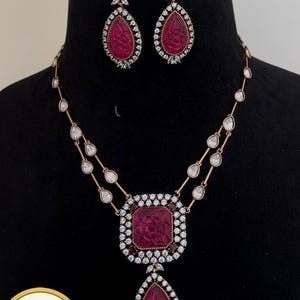 Könnte beinhalten: Ein handgefertigtes Schmuckset mit Halskette und passenden Ohrringen. Das Set umfasst einen quadratischen Anhänger und tropfenförmige Ohrringe mit einem geschnitzten roten Edelstein-Design, umgeben von kleinen weißen Steinen. Die Halskette und Ohrringe sind in goldfarbenem Metall gefasst.