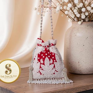 May include: A red and white beaded drawstring bag with a pearl handle. The bag features intricate white floral patterns and a beaded fringe. The bag is displayed on a wooden surface next to a vase with white flowers. The bag is labeled "Daily Designs By Swati Indian Jewelry."