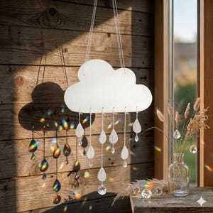Op de afbeelding: Een witte, wolk-vormige hangdecoratie met druppelvormige elementen en heldere kristallen kralen. Het licht breekt door de kristallen en creëert een regenboogeffect op een houten wand.