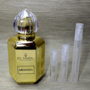 Op de afbeelding: Een gouden fles Medania-parfum van El Nabil, een luxe parfummerk. De fles heeft een doorzichtige glazen dop met een decoratief ontwerp. Drie doorzichtige glazen proefmonsters met spuitkoppen zijn ook afgebeeld.
