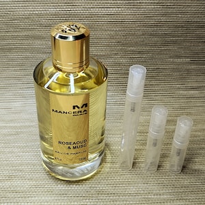Op de afbeelding: Een fles van 120 ml Mancera Paris Rose Aoud & Musc Eau de Parfum. De fles is goudkleurig met een gouden dop en een heldere vloeistof erin. De fles staat op een bruin oppervlak met drie kleine, doorzichtige glazen proefbuisjes.