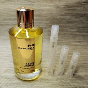 May include: A clear glass bottle of Mancera Roses Jasmine Eau de Parfum, a 4 fl oz / 120 ml bottle with a gold cap and label. The bottle is sitting on a brown textured surface with four clear glass sample vials.