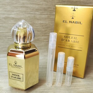 Puede incluir: Una botella dorada de perfume El Nabil Soleil D'Arabie con un tapón de vidrio transparente. La botella está sobre una superficie marrón junto a tres pequeñas botellas de muestra del mismo perfume.